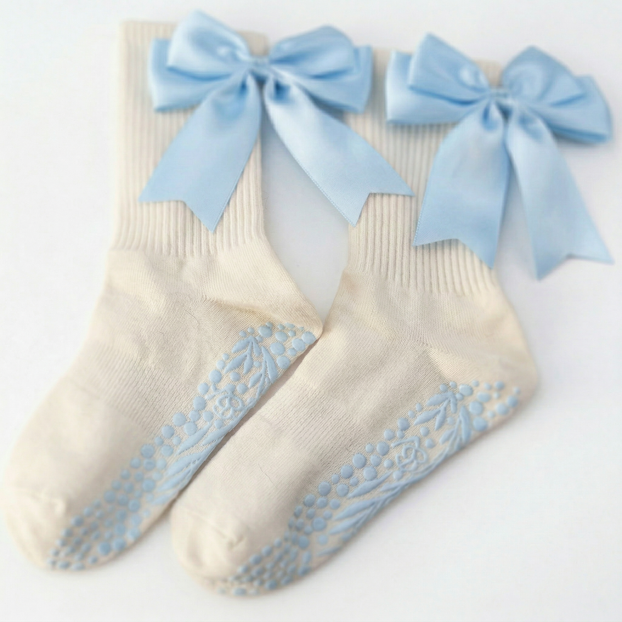 The Signature Ribbon Pilates Socks – Organic Cotton - Baby Blue are cream socks with blue high-traction grips and large, vintage-inspired light blue satin bows at the top.
