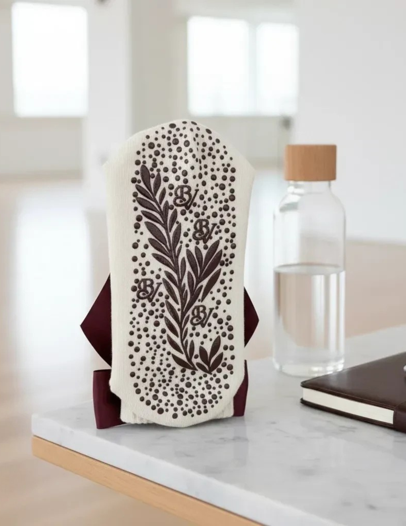 A decorative, cream-colored object with dark brown floral and dotted patterns sits upright on a table next to a glass water bottle with a wooden lid and a closed dark notebook in a bright, modern room.