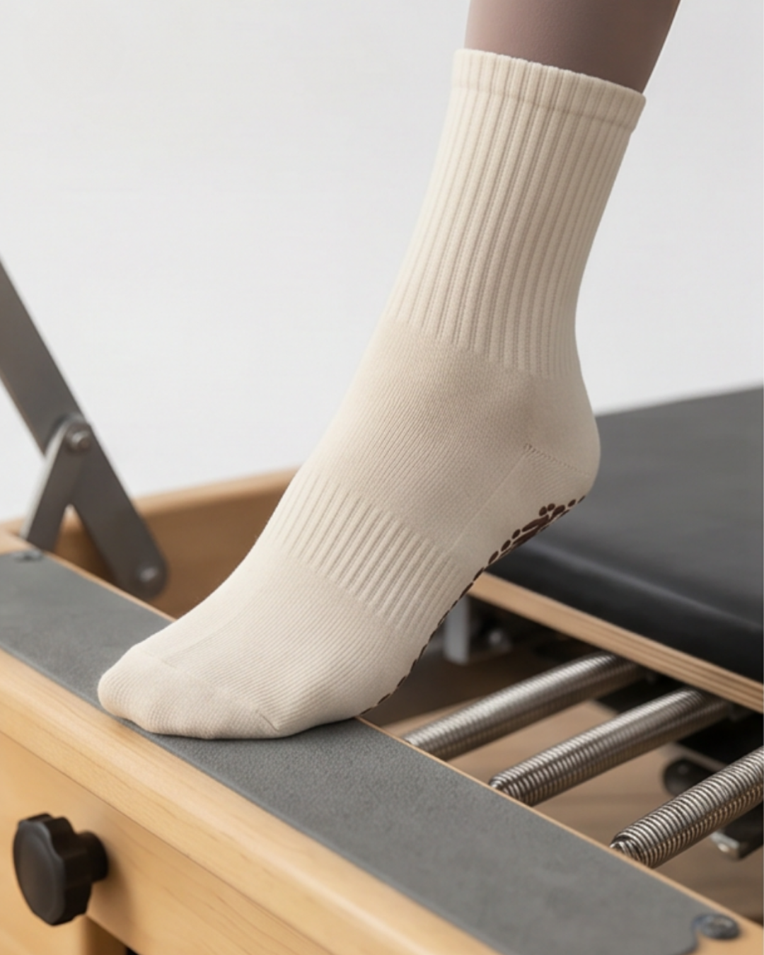 A person wearing The Minimalist Grip Crew – Organic Cotton socks in brown, featuring grip dots on the sole, stands on a wooden Pilates reformer against a neutral background.
