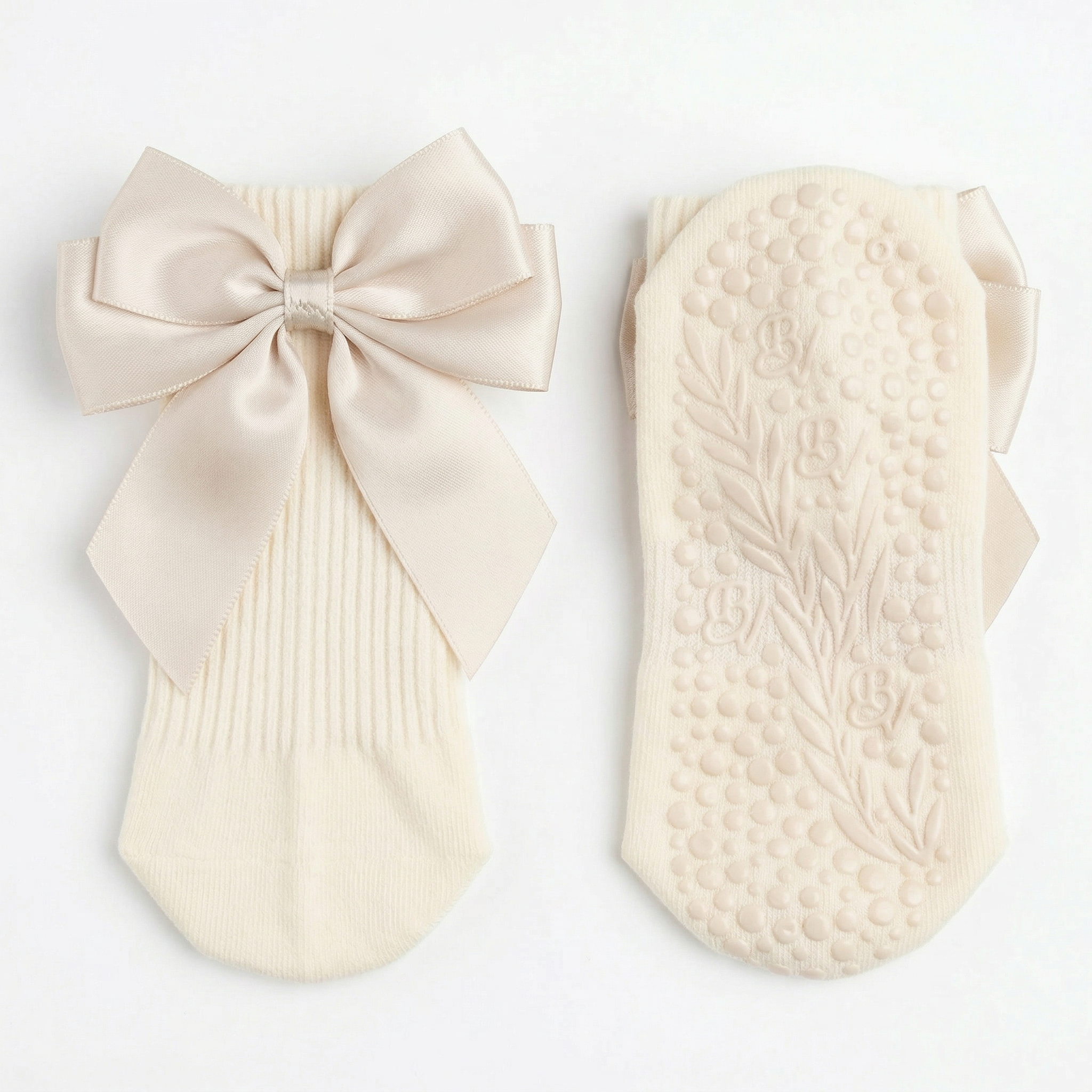 The Signature Ribbon Pilates Socks in light beige, made from organic cotton—one with a vintage-style satin bow on the back, the other with high-traction grip and a leaf pattern on the sole—are displayed against a white background.
